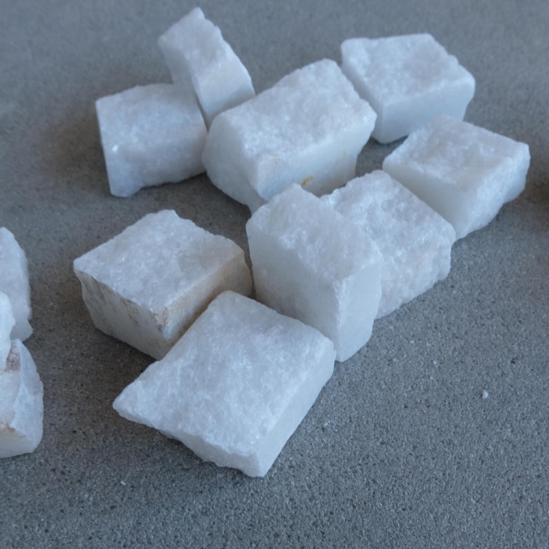 May include: A collection of white, roughly-cut quartz stones. The stones are rectangular and have a slightly rough texture. The stones are scattered on a gray surface. The quartz stones are a bright white color.