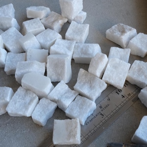 May include: A collection of small, white, irregularly shaped marble tiles. The tiles are scattered across a surface, with a stainless steel ruler providing scale. The tiles appear to be approximately 1 inch in size, suitable for mosaic projects or decorative use.