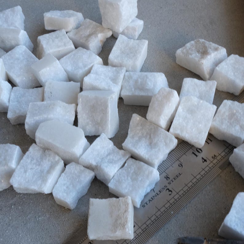 May include: A collection of small, white, irregularly shaped marble tiles. The tiles are scattered across a surface, with a stainless steel ruler providing scale. The tiles appear to be approximately 1 inch in size, suitable for mosaic projects or decorative use.