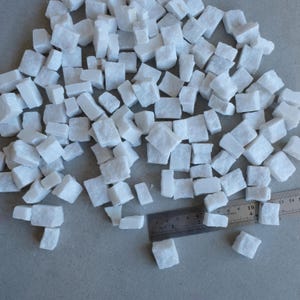 May include: A collection of small, white, irregularly shaped marble cubes. A metal ruler is included for scale, showing the cubes are approximately 0.5 to 1 inch in size. The cubes are scattered on a light gray surface.
