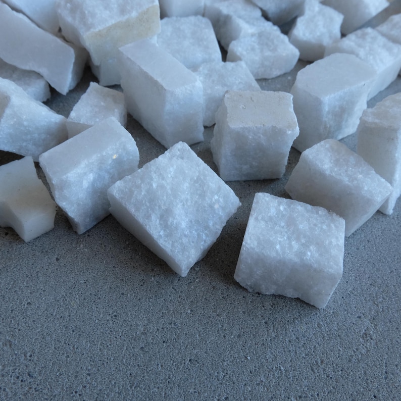 May include: A collection of irregularly shaped, white marble stones. The stones vary in size and shape, with some appearing as small cubes and others as rectangular blocks. The marble has a slightly rough texture and sits on a gray surface.