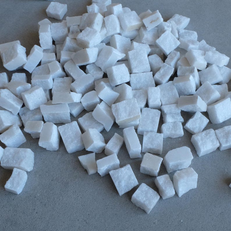 May include: A pile of small, irregularly shaped white marble cubes. The cubes are scattered across a light gray surface, with varying sizes and textures. The overall appearance is clean and natural, suitable for decorative purposes.