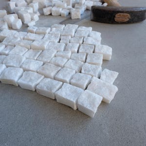 May include: A collection of small, white, square-shaped marble tiles arranged on a gray surface. A hammer rests in the background, suggesting the tiles are being prepared for installation. The tiles are of varying sizes and shapes.