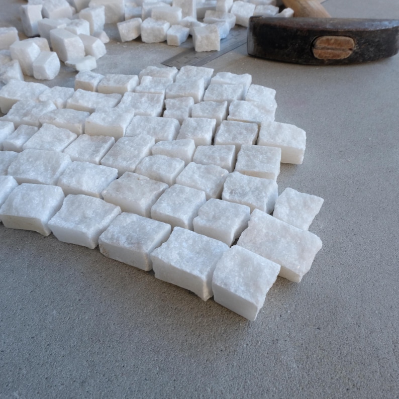May include: A collection of small, white, square-shaped marble tiles arranged on a gray surface. A hammer rests in the background, suggesting the tiles are being prepared for installation. The tiles are of varying sizes and shapes.