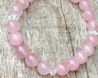 May include: A beaded bracelet featuring large, round, light pink rose quartz beads. The bracelet is interspersed with small, clear, faceted crystal beads. The bracelet is displayed on a weathered, light brown wooden surface.