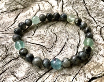 May include: A beaded bracelet featuring a mix of gray and blue-green gemstone beads. The bracelet includes a variety of bead sizes and colors, with a mix of gray, blue, and green hues. The bracelet is displayed on a textured wooden surface.