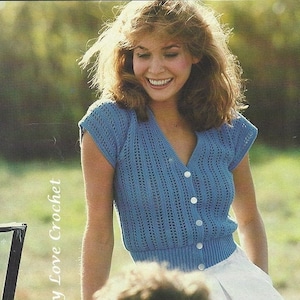 以下が含まれることがあります： Vネックと短い袖の青いクロシェトップ。トップはボタン留めと白い背景になっています。画像の左側には「Sassy Love Crochet」というテキストが表示されています。