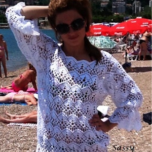 以下が含まれることがあります： 白いクロシェのビーチカバーアップ。貝殻の形のネックラインと長袖。繊細なオープンステッチパターンでできています。画像の右下隅に「Sassy Love Crochet」というテキストが表示されています。