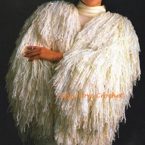 以下が含まれることがあります： クリーム色の、長いフリンジが付いたシャギーかぎ針編みジャケット。 ジャケットはVネックのデザインで、テクスチャード加工の糸で作られています。 画像には「Sassy Love Crochet」の文字が見えます。 ジャケットはクリーム色のタートルネックの上に着用されています。