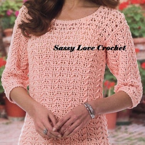 May include: A light peach-colored crochet top with three-quarter sleeves. The top features a textured stitch pattern and a round neckline. The text "Sassy Love Crochet" is displayed across the front of the top. The top is a handmade garment.
