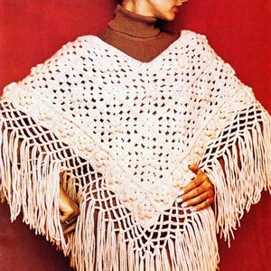 May include: Cream-colored crochet poncho with a V-neck and fringe. The poncho features a textured pattern and long fringe along the bottom edge. The garment is displayed against a red background.