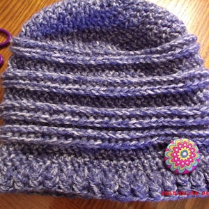 Puede incluir: Un gorro de punto morado y blanco con un botón de flor colorido. El gorro está hecho con un punto texturizado y tiene un borde doblado.