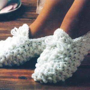 May include: A pair of white, textured, crocheted slippers. The slippers have a rounded toe and a small bow detail at the top. The slippers are on a wooden surface.