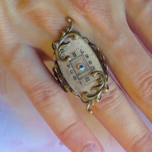 May include: A vintage-style ring with a white oval face featuring a clock face with Roman numerals and a blue jewel in the center. The ring is made of brass with ornate scrollwork.