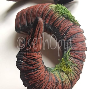 May include: Two curved, horn-shaped decorative pieces. The horns are primarily reddish-brown with black accents, textured to resemble wood grain. Green moss adds detail. The artist's signature, "sethOpia", is visible.