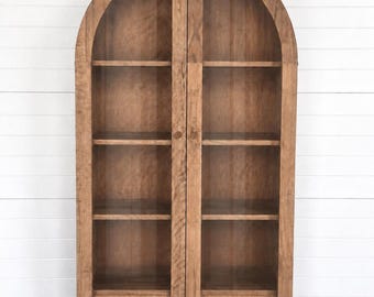 May include: A tall, arched wooden cabinet featuring two glass-paneled doors, four internal shelves, and two bottom drawers with circular wooden knobs. The cabinet is constructed from light brown wood with a visible grain and stands on four short legs.