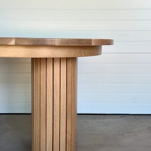 May include: A close-up of a wooden oval table with a slatted base. The table top is light wood and the base is a darker wood.