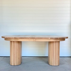 May include: A light wood oval dining table with a unique slatted base. The table is made of solid wood and has a natural finish.