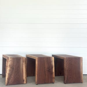 May include: Three handcrafted wooden side tables with a natural edge. The tables are made of dark brown wood with visible grain and a rectangular shape. The tables are placed on a concrete surface against a white wall.
