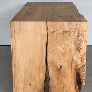 May include: A solid wood table with a natural edge and a light brown finish. The table has a rectangular top and a thick, block-like base. The wood grain is visible throughout the piece, adding to its rustic charm.