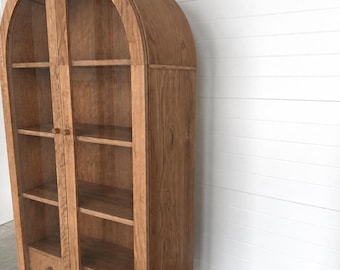 May include: A tall, arched wooden cabinet featuring two glass-paneled doors with small round knobs. The interior contains four horizontal shelves, and the base includes two drawers with matching round wooden knobs, all supported by four short legs.