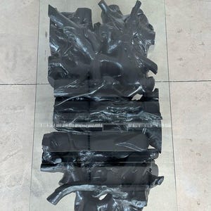 May include: A modern coffee table with a clear glass top and a dark gray, sculptural base. The base appears to be made of carved wood or a similar material, with an abstract, organic design. The table is set on a light-colored tiled floor.