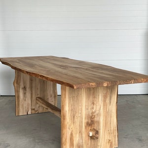 May include: A large, rustic wooden dining table with a live edge top and a unique, asymmetrical base. The table is made of light brown wood with a natural finish.