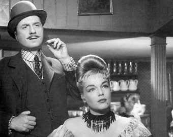 May include: A black and white photo of a man and a woman in a vintage setting. The man is wearing a suit and a bowler hat. The woman is wearing a dress with a lace collar and a beaded necklace.