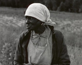 May include: A black and white photo of an older woman wearing a white headscarf, a striped shirt, and a gingham apron. She is holding a wooden stick and looking off to the side.