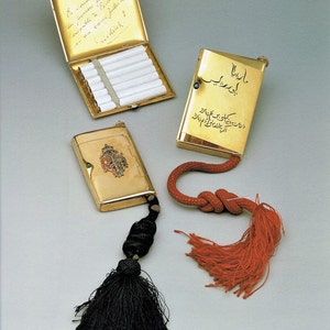Puede incluir: Tres estuches de cigarrillos de oro con borlas. El estuche de la izquierda tiene una inscripción manuscrita en francés. El estuche del centro tiene una inscripción en árabe. El estuche de la derecha tiene un pequeño diseño colorido.