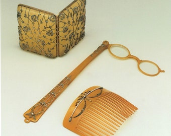 Op de afbeelding: Een set van drie antieke goudkleurige accessoires: een klein, scharnierend, met juwelen bezet compact, een vergrootglas op een lange steel en een kam met een juweelontwerp.