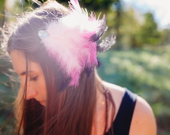May include: A woman with long brown hair wearing a pink and black feather headband with a sparkly jewel accent. The headband is worn on the side of her head. The background is out of focus and appears to be a green grassy field.