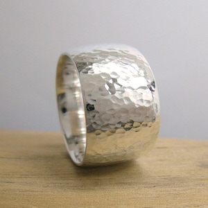 May include: A wide silver band ring with a hammered texture. The ring is made of sterling silver and has a polished finish.