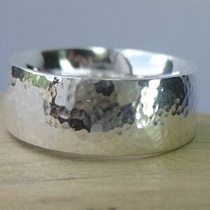 Peut inclure: Une large bague en argent martelé. La bague a une surface texturée avec une série de petites indentations irrégulières. La bague est de couleur argent brillant.