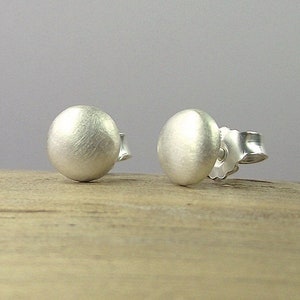 May include: Two silver stud earrings with a matte finish. Each earring is a small, rounded sphere. The earrings are displayed on a light brown wooden surface against a gray background. The earrings have a post and butterfly backing.