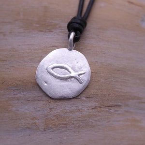 Pode incluir: Um colar com um pingente de prata e um cordão de couro preto. O pingente é um disco redondo com um design de peixe estilizado.