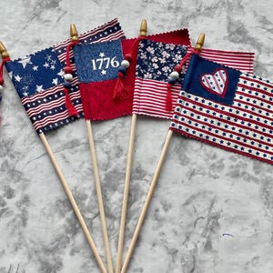 May include: Five small American flags with wooden dowels. The flags feature patriotic designs, including stars, stripes, and floral patterns. One flag displays the number "1776". Each flag is adorned with decorative tassels and beads.