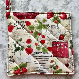 May include: Quilted square pot holder with a strawberry theme. The off-white fabric features red strawberries, green leaves, and white blossoms. Text includes "Feeling Berry Good" and "Strawberries, Always Sweet & Juicy."