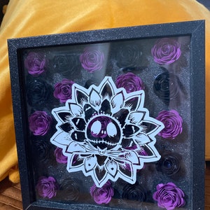 May include: A black shadow box frame with a white and black Jack Skellington design in the center. The frame is filled with purple and black paper flowers.