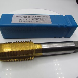 Può includere: Un maschio per filetti a mano dorato con una custodia in plastica blu. La custodia ha un'etichetta che recita "MADE PERFECT HAND TAPS & TOOL COMPANY M30 X 3.5 H2 HSS BOTOMING TAP III".