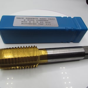 Può includere: Un maschio per filetti a mano dorato con una custodia in plastica blu. La custodia ha un'etichetta che recita "MADE PERFECT HAND TAPS & TOOL COMPANY M30 X 3.5 H2 HSS BOTOMING TAP III".