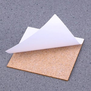May include: A square cork sheet with a white adhesive backing. The backing is partially peeled back to reveal the adhesive.