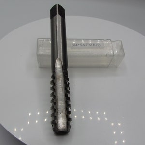 Può includere: Un maschio in metallo argentato con un tagliente scanalato e un'estremità quadrata. Il maschio è posizionato verticalmente. Dietro il maschio c'è un astuccio di plastica trasparente con testo.