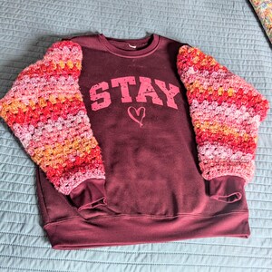 May include: A maroon sweatshirt featuring the word STAY in distressed pink lettering above a small pink heart graphic. The long sleeves are made of multi-colored crocheted yarn in shades of pink, orange, and red with ribbed cuffs and hem.