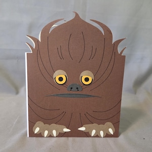 May include: A brown papercraft card depicting a cartoon Wookiee character. The card has a square shape with a detailed face, including yellow eyes, a gray nose, and a mouth. The card also has paws. The card is made of paper.