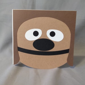 May include: A brown paper card with a cut-out of a dog's face. The dog has white eyes, a black nose, and a black mouth.
