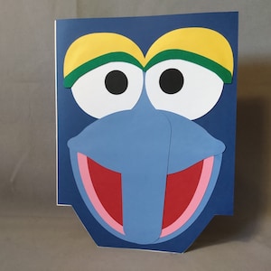May include: A blue paper craft of a character with yellow eyebrows, green eyelids, white eyes, a blue nose, and a red mouth.