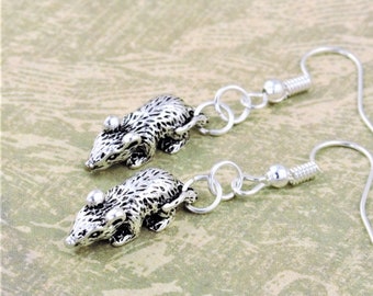 May include: A pair of silver dangle earrings featuring a small, detailed animal charm. The charm resembles a mouse or a small rodent with a textured surface. The earrings have a simple hook closure.
