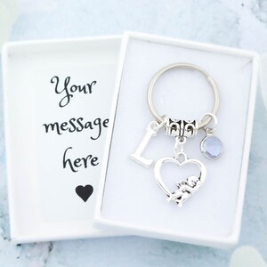 以下が含まれることがあります： ハート型のチャームにキューピッドが描かれたシルバーのキーホルダー。Lの文字のチャームとライトブルーのクリスタルチャームも付いています。キーホルダーは、白い箱に入っていて、「メッセージはこちら」という文字と小さな黒いハートが描かれています。