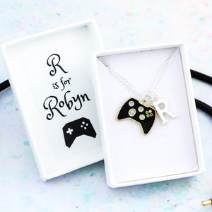 May include: A white box with a black and white illustration of a video game controller and the text "R is for Robyn". A silver chain necklace with a black video game controller charm and a silver letter "R" charm is in a white box.